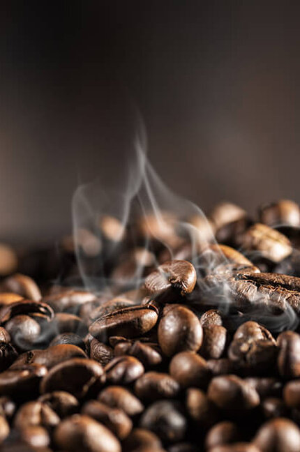 A close-up of freshly roasted coffee beans with wisps of steam rising, set against a dark background.