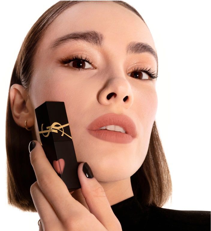 A close-up of a woman with a sleek hairstyle holding a lipstick tube decorated with a gold ribbon and a heart design, wearing neutral makeup and posing against a white background.