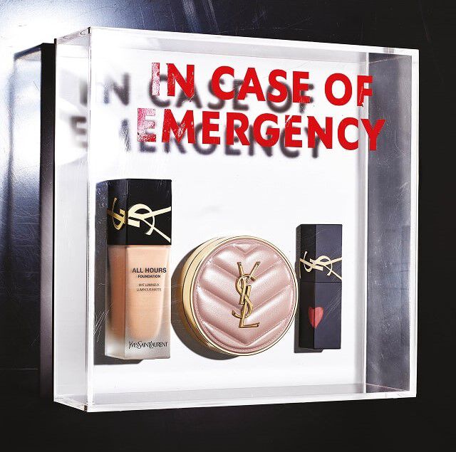 Una scatola trasparente in plexiglas contiene tre prodotti di make-up YSL Beauty, come un kit di emergenza. All'interno, da sinistra a destra, si vedono un flacone di fondotinta liquido "All Hours Foundation", una cipria compatta o un cushion colorato in una custodia trapuntata rosa pallido con il logo YSL dorato, e un rossetto nero decorato con un piccolo cuore rosso sul tappo, anch'esso con il logo YSL dorato. Sul retro della scatola trasparente, il testo "IN CASE OF EMERGENCY" e scritto in grandi lettere rosse, creando un effetto specchio e di profondita, suggerendo che questi prodotti sono essenziali da avere sempre a portata di mano.