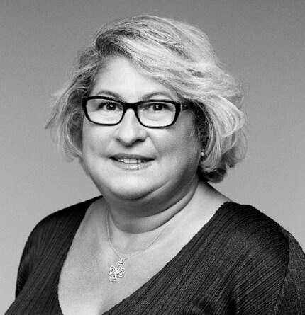 A smiling woman with curly hair wearing glasses and a dark top, posed against a neutral background.