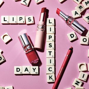 Rossetti YSL e matita labbra su sfondo rosa con lettere dello Scarabeo che formano le parole "LIPS", "LIPSTICK" e "DAY"