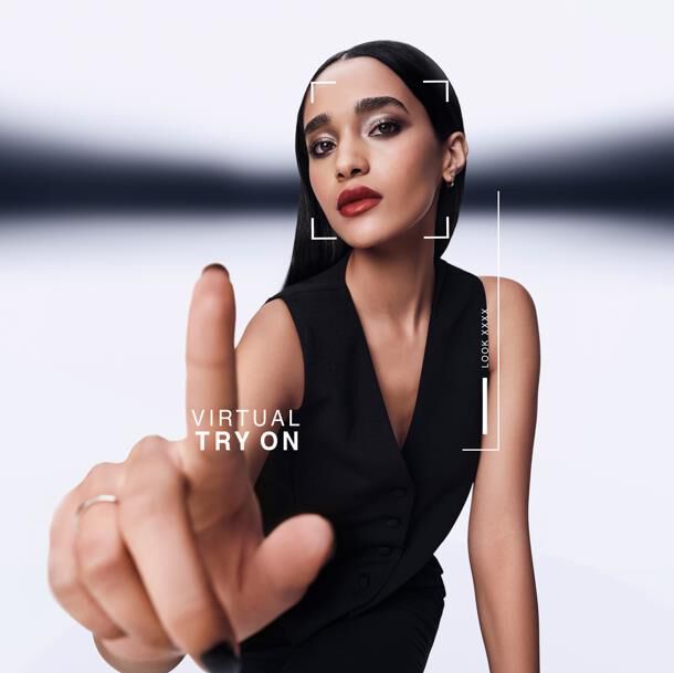 Donna punta in avanti, interagendo con uno schermo di prova virtuale. Grafica mostra "VIRTUAL TRY ON" e "LOOK XXXX" attorno al suo viso. Indossa un gilet nero.​