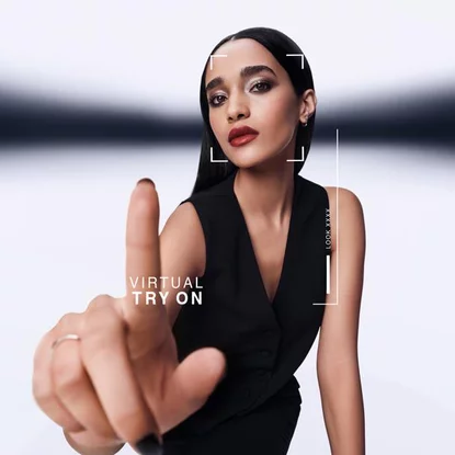 Donna punta in avanti, interagendo con uno schermo di prova virtuale. Grafica mostra "VIRTUAL TRY ON" e "LOOK XXXX" attorno al suo viso. Indossa un gilet nero.
