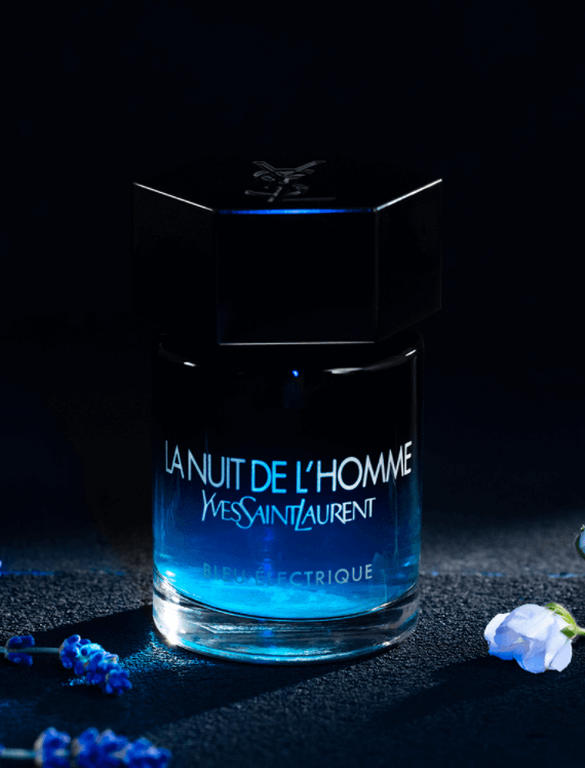 A bottle of "La Nuit de L'Homme" by Yves Saint Laurent, featuring a dark blue gradient and hexagonal black cap, surrounded by lavender and white flowers against a dark background.