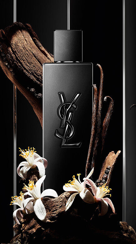 Flacone di profumo YSL nero circondato da legno, baccelli di vaniglia e fiori bianchi.