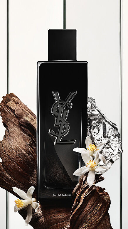 Una bottiglia nera di Eau de Parfum Yves Saint Laurent con il logo YSL, circondata da pezzi di legno scuro, delicati fiori bianchi e un oggetto riflettente argentato stropicciato.