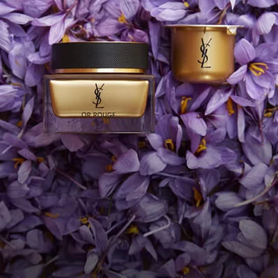A gold and black YSL skincare jar and a small container are placed on a bed of purple petals.
