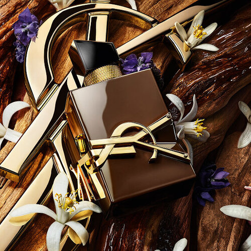 Flacone di profumo YSL Libre circondato da dettagli dorati, fiori d'arancio bianchi e fiori viola su sfondo di legno.