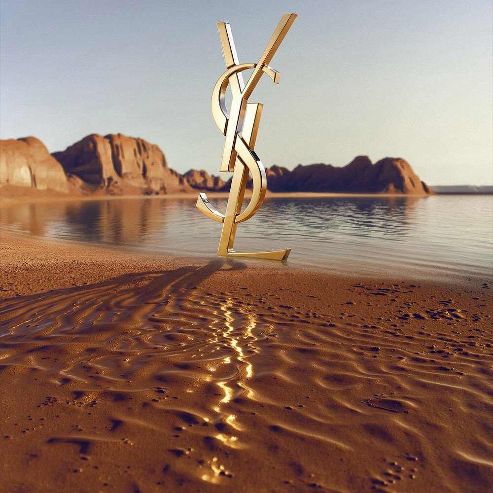 Un grande logo dorato YSL (Yves Saint Laurent) in 3D su una costa desertica. Il monogramma in oro lucido riflette la luce del sole ed e posizionato davanti a uno specchio d'acqua calmo con spettacolari formazioni rocciose rosse e montagne desertiche sullo sfondo sotto un cielo sereno.