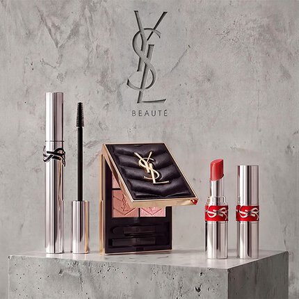 Presentazione promozionale di prodotti di trucco YSL Beaute tra cui mascara, una palette di ombretti trapuntata e rossetti su sfondo in cemento.