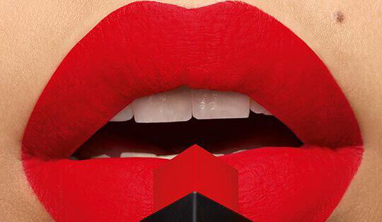Close-up of a pair of lips painted bright red, with a red lipstick tube positioned at the center. The shiny finish contrasts with the matte texture of the lips.