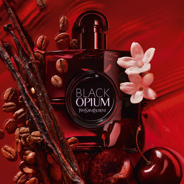A bottle of Black Opium perfume by Yves Saint Laurent, surrounded by coffee beans, red cherries, and white flowers against a rich red background.