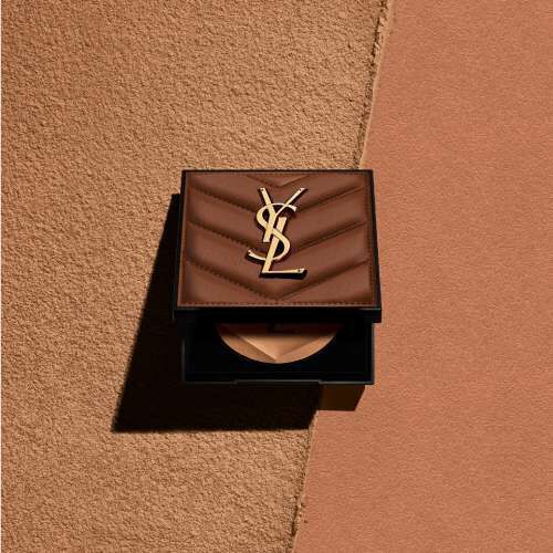 A compact makeup case featuring the classic YSL logo, displayed on a textured beige background that transitions to a warm brown surface.