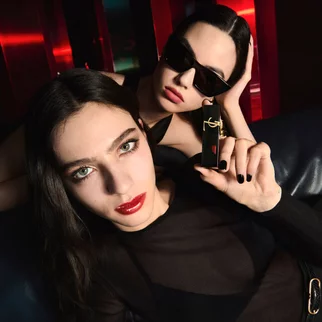 Two individuals with dark hair and bold makeup pose for a beauty shot. One holds a lipstick product, showcasing a striking red lip, while the other leans back stylishly, wearing sunglasses. The background features vibrant, colorful lighting.