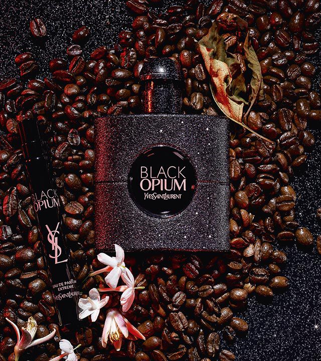 A black bottle of Yves Saint Laurent's Black Opium perfume surrounded by coffee beans and white flowers, with a smaller rollerball fragrance beside it.