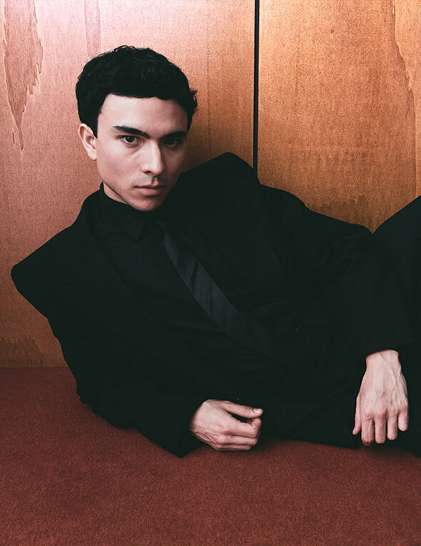 Un giovane uomo in abito elegante nero, sdraiato contro uno sfondo di legno chiaro, guarda l'obiettivo con un'espressione intensa.