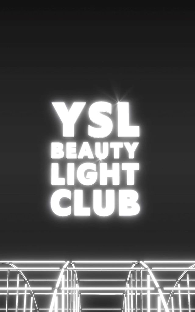 Un'ampia e scura inquadratura rivela il logo "YSL BEAUTY LIGHT CLUB" in lettere bianche luminose, fluttuante al centro di uno spazio nero. Sotto il logo, una complessa struttura di barre metalliche o neon bianchi luminosi crea un effetto griglia o palcoscenico, suggerendo un'installazione luminosa o un ambiente da club futuristico. L'insieme emana un'atmosfera moderna ed elegante.
