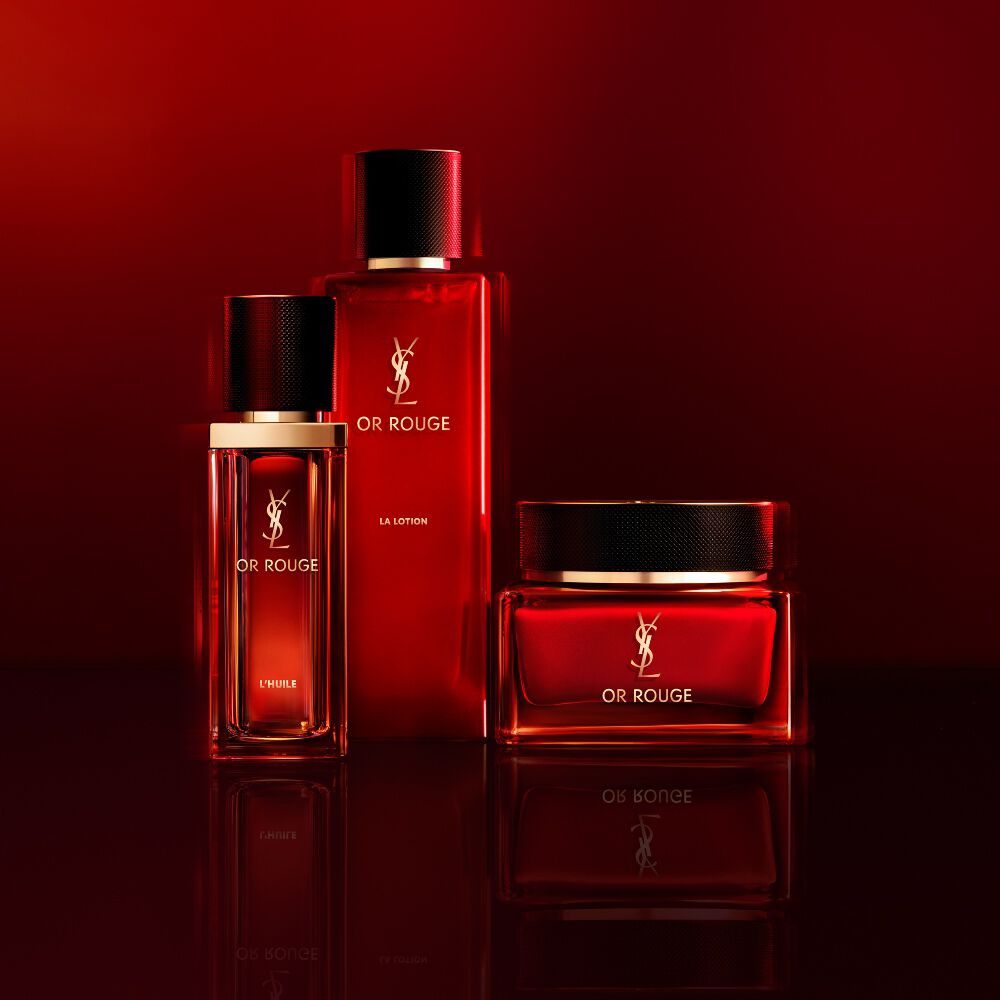 Tre prodotti per la cura della pelle Yves Saint Laurent Or Rouge in flaconi e vasetto di vetro rosso traslucido su sfondo rosso scuro.