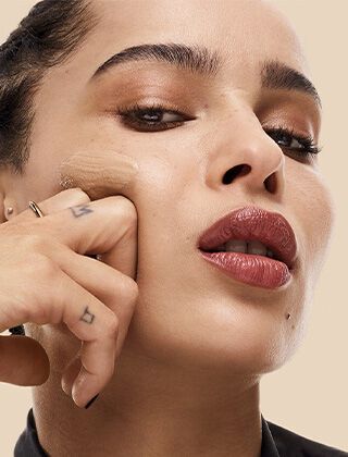 A close-up portrait of a woman applying foundation to her face with a finger, showcasing a confident expression and glossy lips against a light beige background.