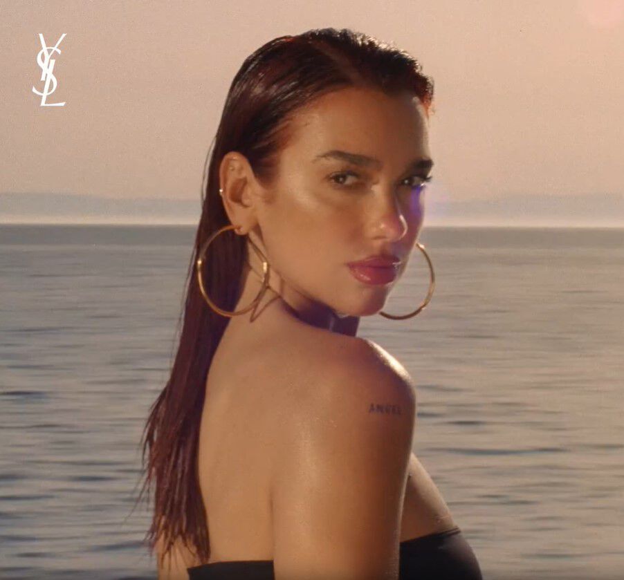 A woman with long, wet hair gazes over her shoulder at the camera, set against a serene ocean backdrop. She wears large hoop earrings and has a tattoo on her arm. The image has a warm, golden hue, suggesting a sunset atmosphere. The YSL logo is visible in the top left corner.