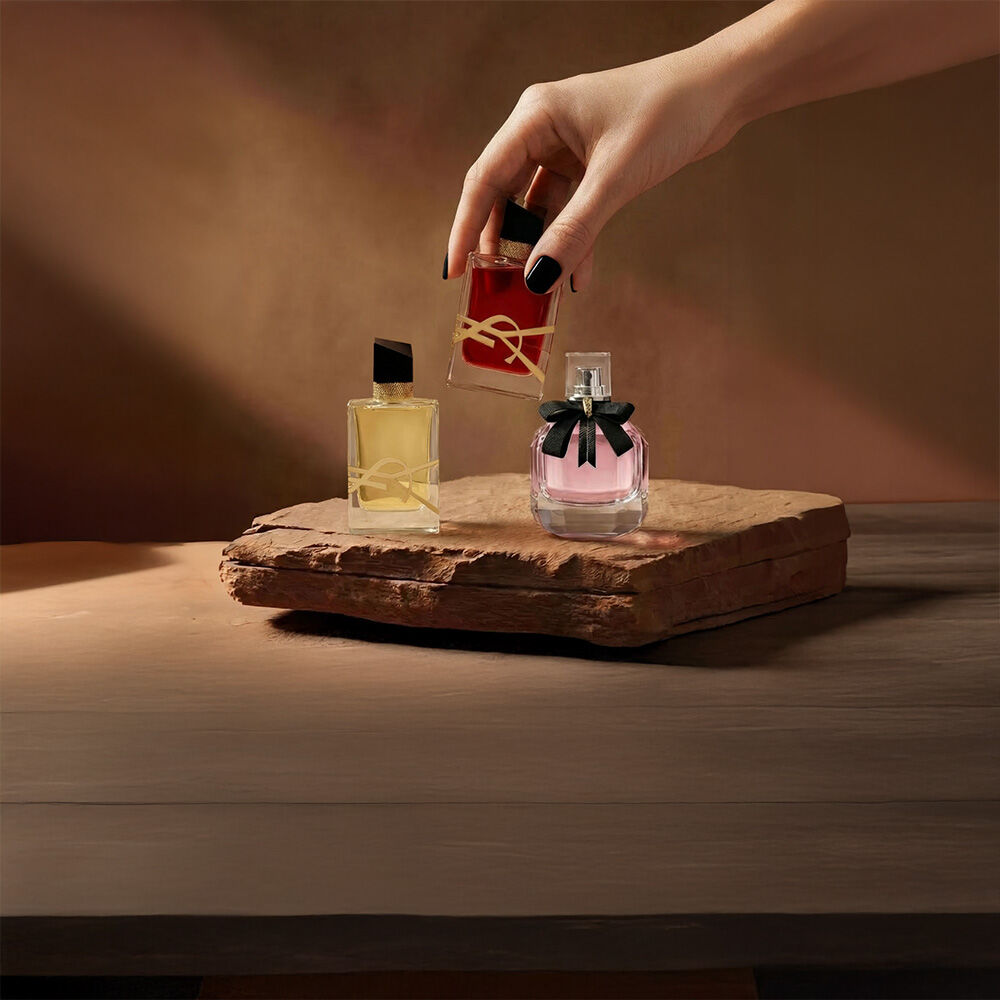 Una mano tiene un flacone di profumo YSL rosso accanto ad altre due fragranze su una base di pietra.