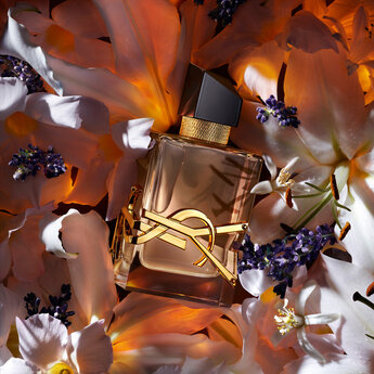 A luxurious perfume bottle is placed among delicate white and purple flowers, illuminated by warm light. The bottle features a gold logo and a sleek design, adding to the elegant aesthetic of the arrangement.