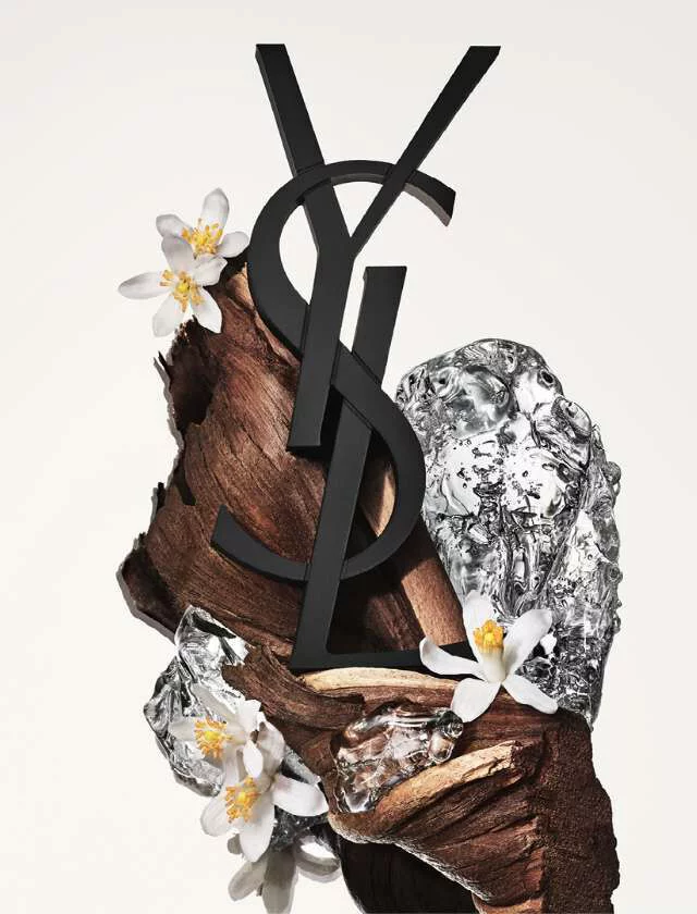 A creative arrangement featuring the YSL logo, intertwined with textured wood, shiny metallic elements, and delicate white flowers, set against a soft, neutral background.