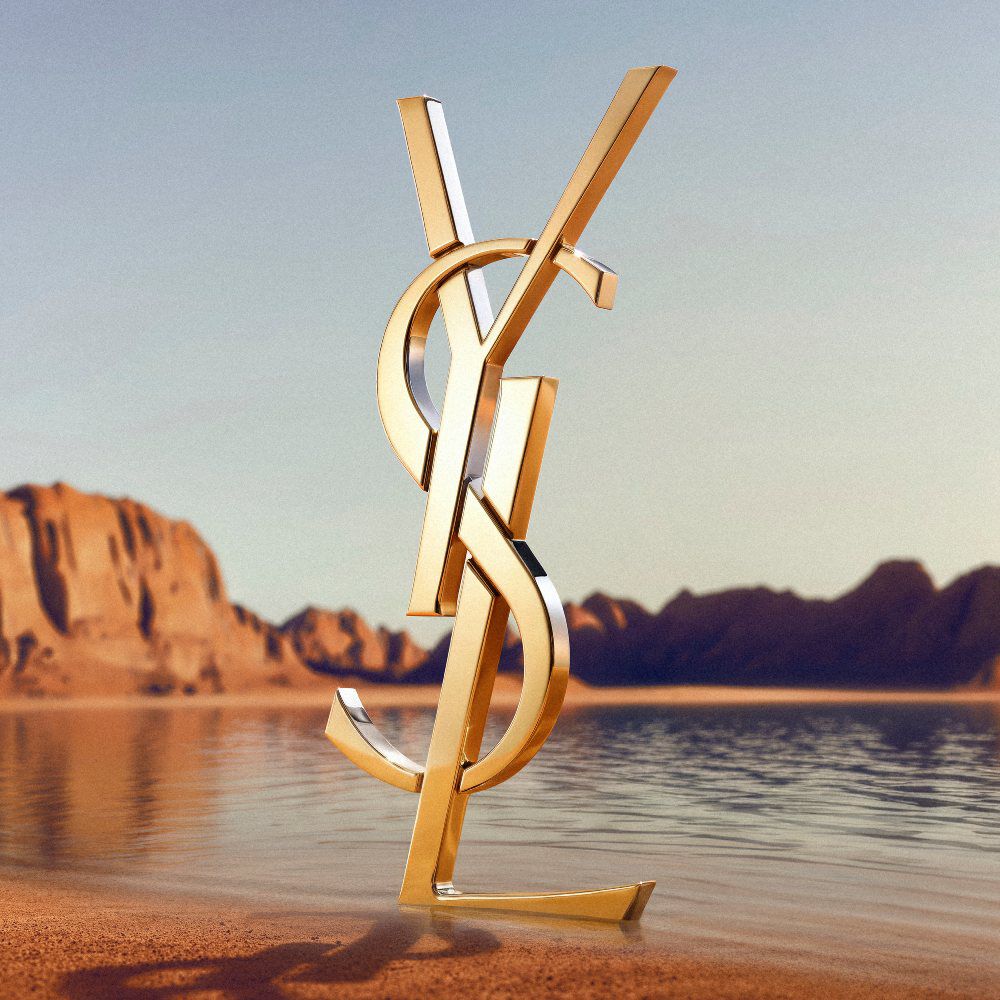 Un grande logo dorato YSL (Yves Saint Laurent) in 3D su una costa desertica. Il monogramma in oro lucido riflette la luce del sole ed e posizionato davanti a uno specchio d'acqua calmo con spettacolari formazioni rocciose rosse e montagne desertiche sullo sfondo sotto un cielo sereno.