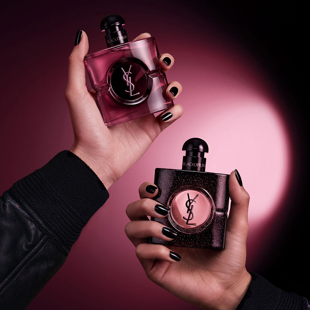 Due mani che reggono flaconi di profumo Yves Saint Laurent Black Opium su uno sfondo rosa scuro e nero.