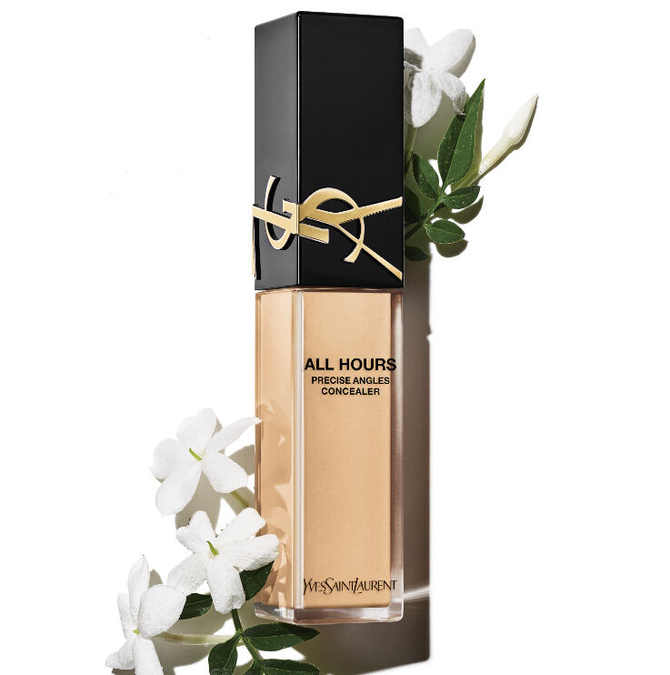A bottle of Yves Saint Laurent All Hours Precise Angles Concealer sits against a white background, surrounded by delicate jasmine flowers. The concealer bottle has a sleek, modern design with a black cap and gold lettering.