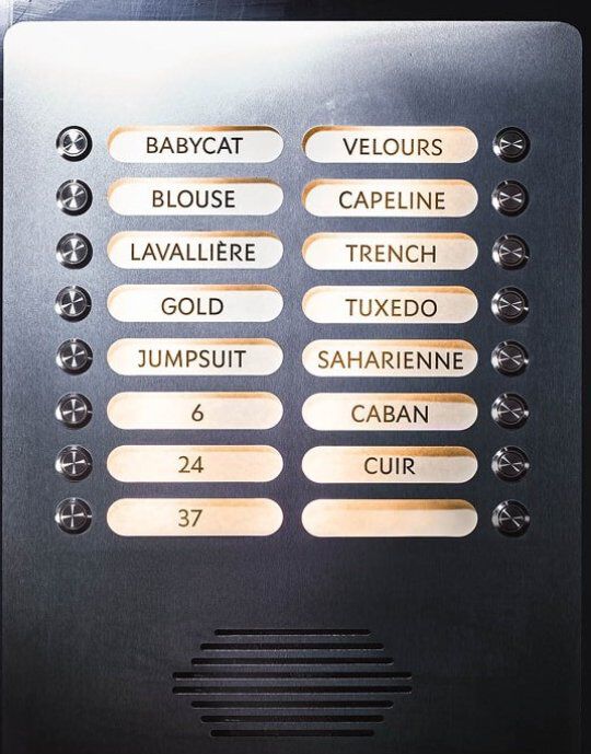 Pannello metallico con pulsanti illuminati, ciascuno etichettato con una parola che si riferisce alla moda YSL. Le etichette includono 'BABYCAT', 'TUXEDO', 'SAHARIENNE' e 'MUSE', tra le altre. Il pannello presenta piccoli pulsanti circolari e una griglia per l'altoparlante in basso, assomigliando a un elegante sistema di interfono.