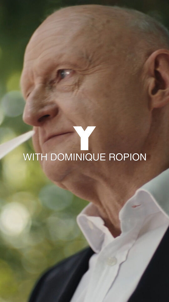 Un uomo calvo in abito nero e camicia bianca tiene al naso diverse strisce di carta bianca, comunemente usate per testare le fragranze, come se stesse valutando un profumo. Ha un'aria contemplativa, con uno sfondo verde e sfocato di fogliame e alberi. L'immagine e sovrastata da un grande testo bianco "Y" e da un testo piu piccolo sotto di esso, "WITH DOMINIQUE ROPION".