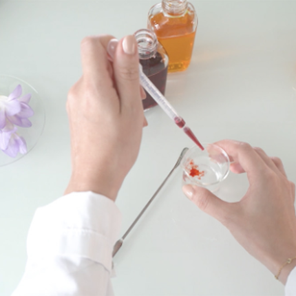 A person holds a pipette over a small dish, adding a red liquid to it, while surrounded by various colored liquids in jars and small cups, along with a few strands of saffron and purple flowers nearby.