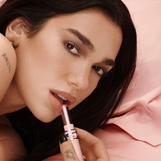 Dua Lipa applica un rossetto YSL su uno sfondo rosa cipria.​