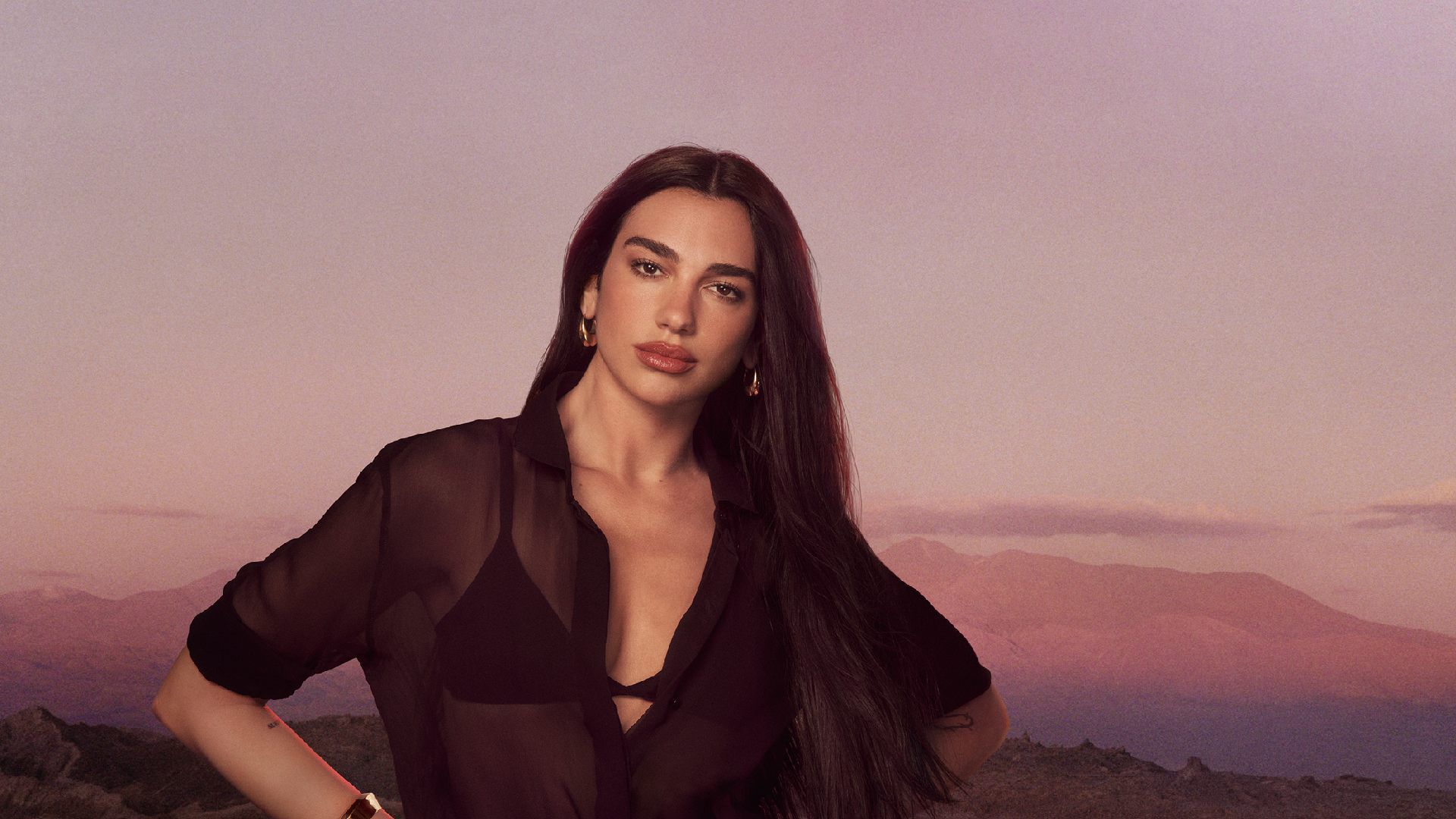 Dua Lipa in posa con una camicia nera trasparente su uno sfondo di montagne al tramonto.