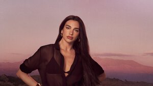 Dua Lipa in posa con una camicia nera trasparente su uno sfondo di montagne al tramonto.