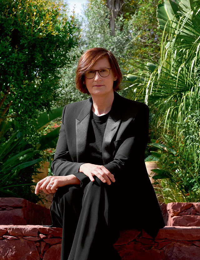 Una donna con capelli castano-rossicci e occhiali siede su un muro di pietra, indossando un abito nero. Sta guardando l'osservatore, con un giardino verde sullo sfondo.