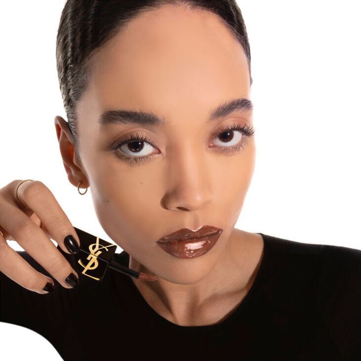 A model with long dark hair and a flawless complexion holds a lip gloss with a bold shade, looking confidently at the camera against a white background.