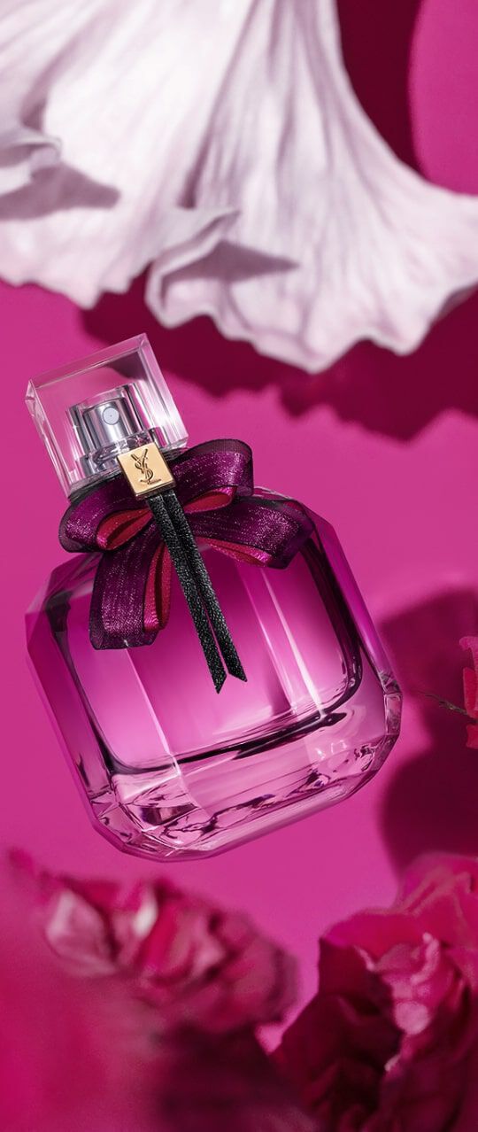 A stylish glass perfume bottle with a decorative purple bow, against a vibrant pink background, surrounded by soft floral elements.