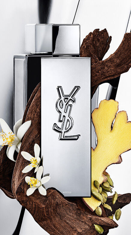 Flacone di profumo YSL Y L'ABSOLU circondato da legno scuro, fiori bianchi di neroli, zenzero e baccelli di cardamomo.