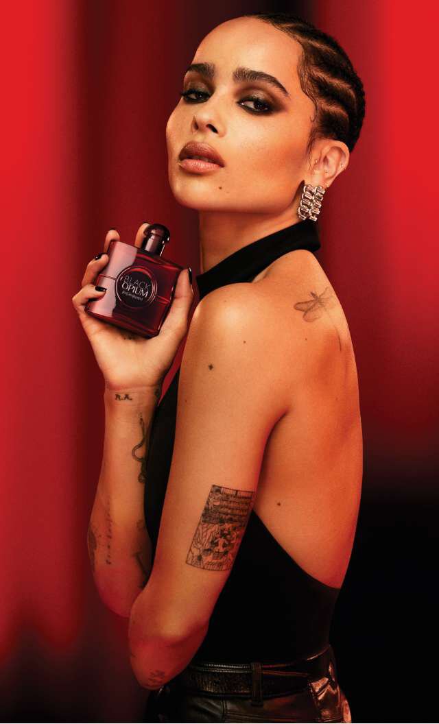 Ritratto di una donna elegante con capelli intrecciati, che tiene in mano una bottiglia di profumo, con uno sfondo rosso.