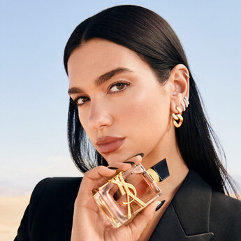 A woman with long, straight dark hair holds a bottle of YSL perfume close to her face, wearing trendy earrings and a sleek black blazer, set against a minimalist background.