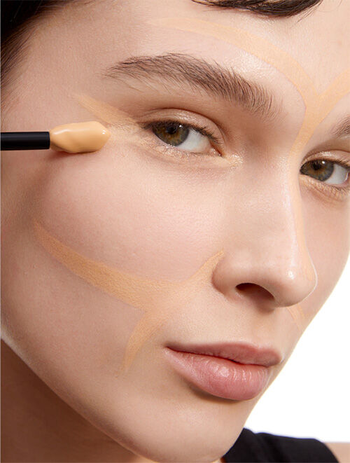 A close-up of a person applying foundation to their face with a makeup brush, showcasing smooth skin and a natural look. The foundation is strategically placed in a pattern on the skin.