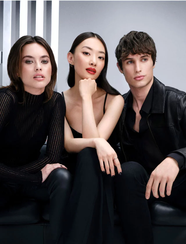 Tre giovani modelli eleganti in abiti neri seduti su un divano in pelle nera su uno sfondo grigio da studio.