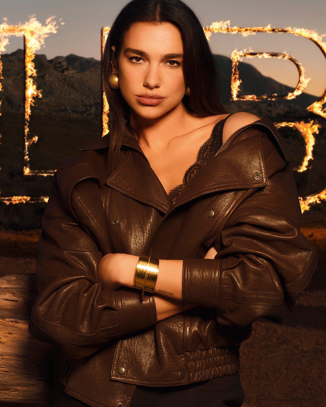 Dua Lipa in una giacca di pelle marrone, con grandi lettere LIBRE in fiamme su un paesaggio desertico e selvaggio.