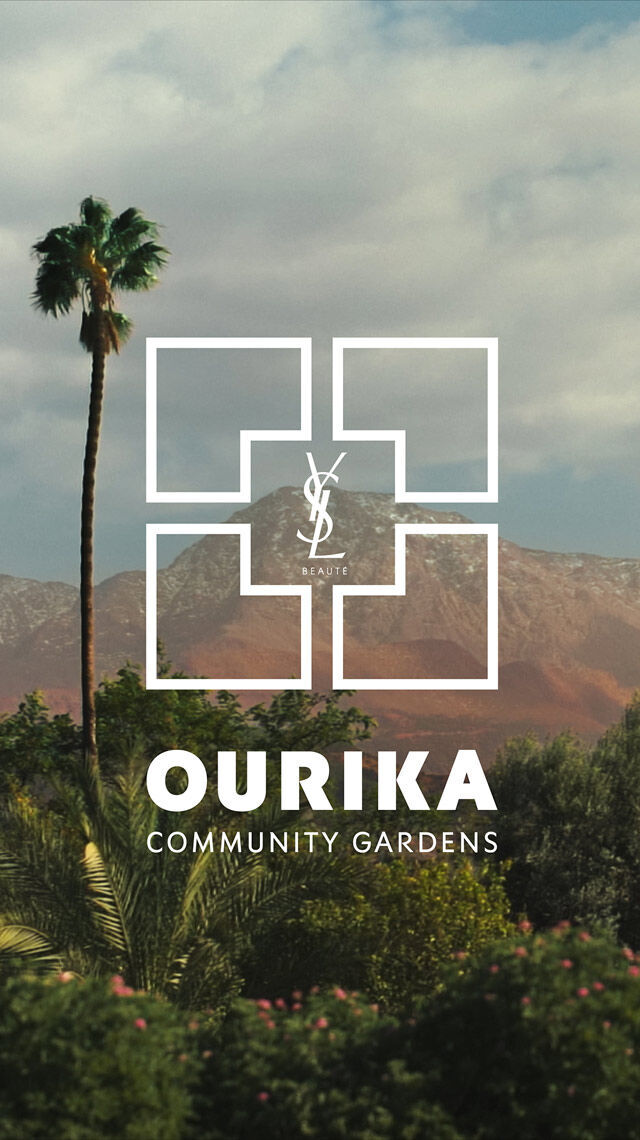 Un paesaggio con palme, fogliame verde e montagne sotto un cielo nuvoloso. Il logo bianco di YSL Beaute e il testo "OURIKA COMMUNITY GARDENS" sono sovrapposti.