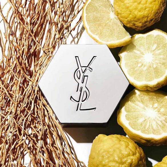 A hexagonal container with the YSL logo is surrounded by halved lemons and rustic, textured materials.