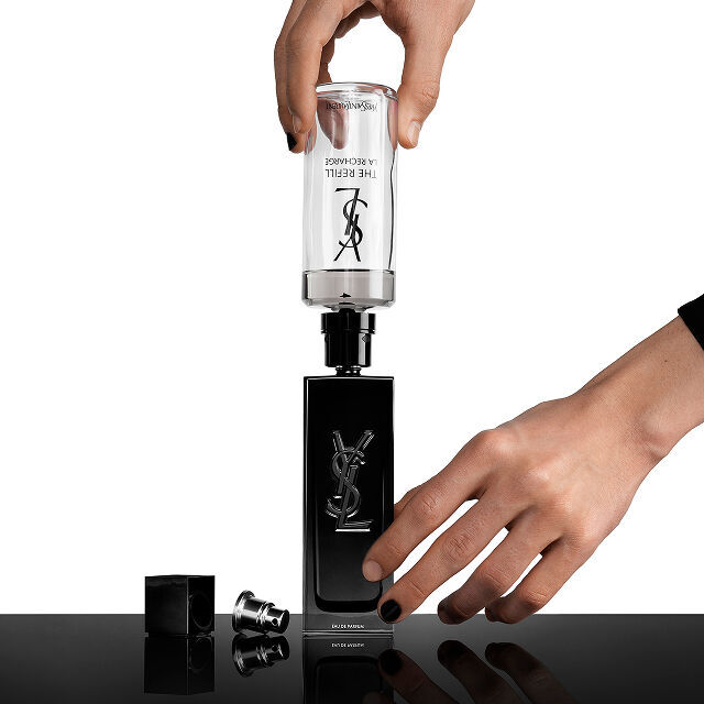 Le mani di una persona ricaricano un flacone di profumo nero Yves Saint Laurent (YSL) con una cartuccia di ricarica trasparente con la scritta "THE REFILL LA RECHARGE."