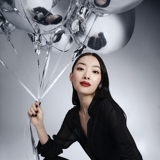 Una donna con lunghi capelli neri, con una camicia nera, tiene in mano dei palloncini argentati e guarda direttamente la fotocamera. Indossa un rossetto rosso.