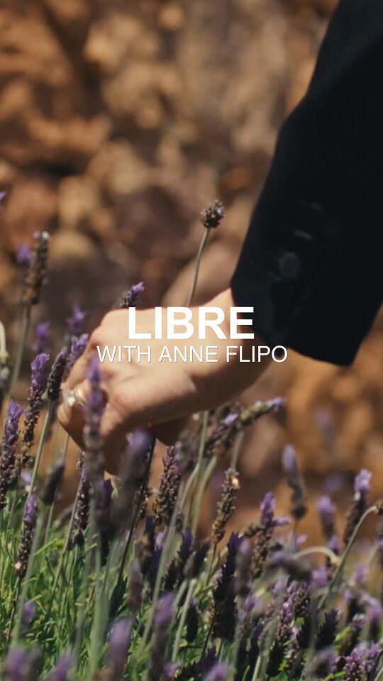 Una mano, forse di Anne Flipo, si protende verso un campo di fiori viola lavanda. La mano indossa un anello ed e parzialmente oscurata dalla scritta in bianco "LIBRE WITH ANNE FLIPO". Sullo sfondo, una parete rocciosa sfocata.